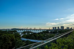 Panoramic view of a coastal cityscape at dusk, featuring a long bridge spanning water with lush green trees in the foreground and modern high-rises in the distance