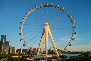 A large white Ferris wheel at dusk seen from below against a clear blue sky, with city towers and open water behind it