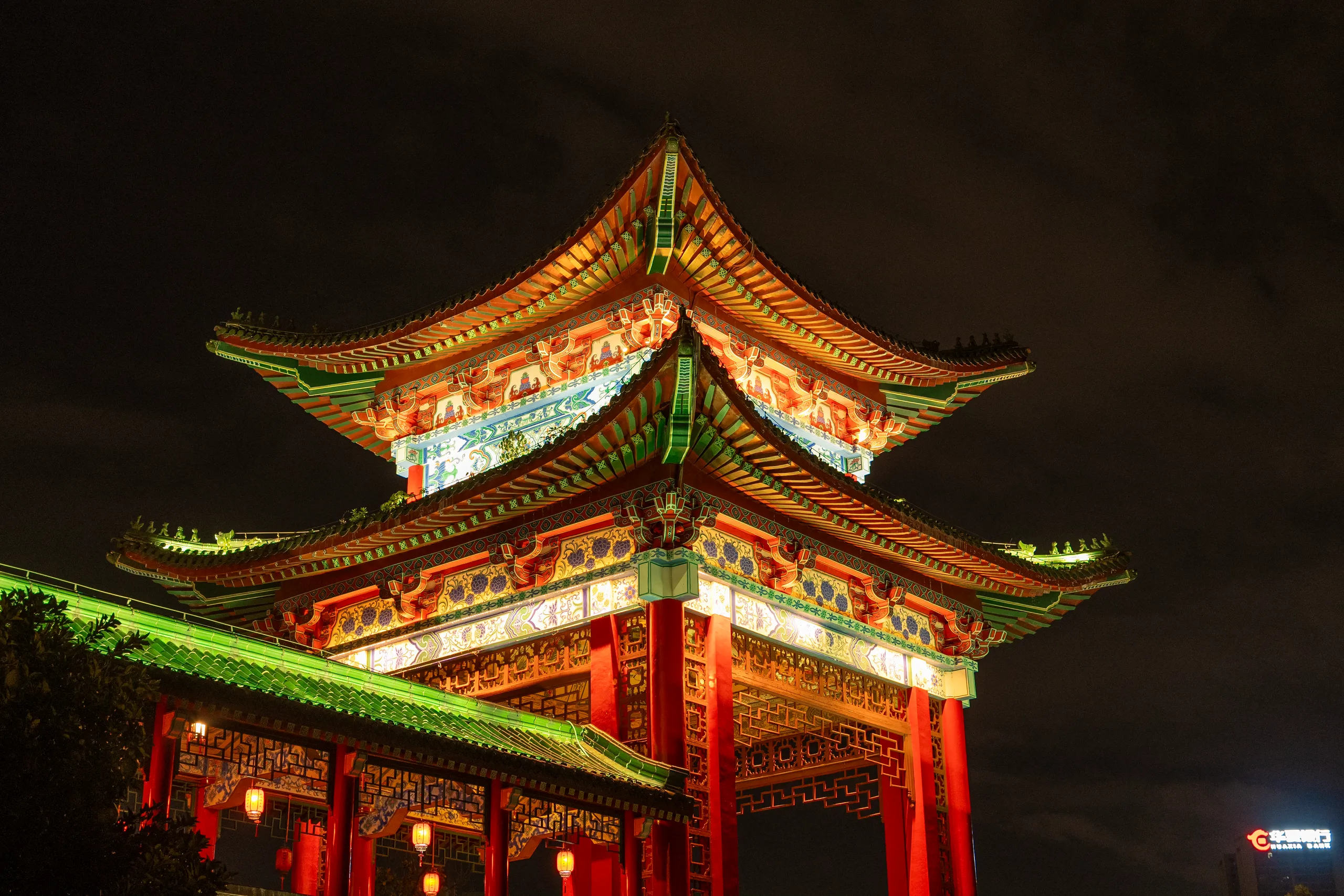 Two-tiered traditional Chinese pavilion with ornate curved roofs and red pillars, illuminated by warm golden and green lighting against dark night sky