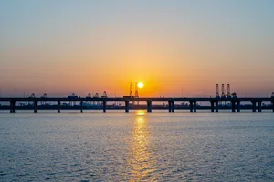 The sun setting directly behind a long low bridge and a row of container-port gantry cranes, laying a column of light across flat water